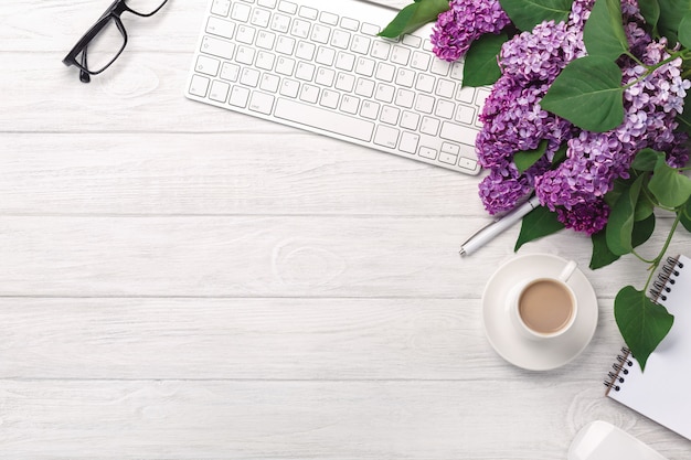 Office desktop with a bouquet of lilacs, coffee cup, keyboard, notebook and pen on white boards