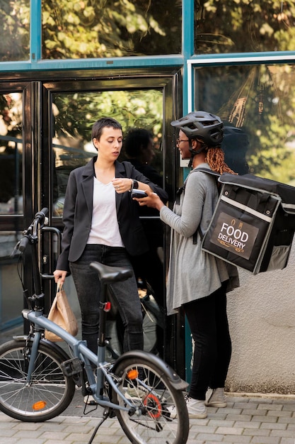 Office employee paying for delivery with smart watch contactless payment transaction. African american courier holding pos terminal, standing near company building outdoors