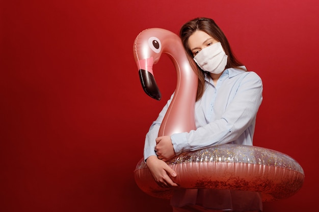 Office employee young beautiful woman on red background in protective medical mask hugs swimming circle pink flamingo in depression, pandemic, vacation cancellation