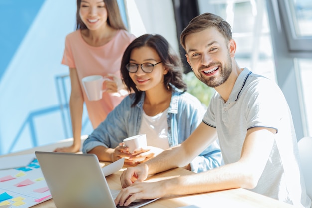 At the office. Happy cheerful delighted man smiling and looking at you while working on a laptop