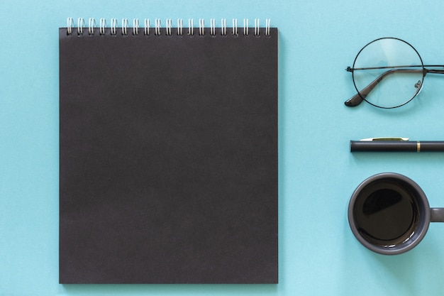 Office or home workplace. Black color notepad, cup of coffee, spectacles, pen on blue