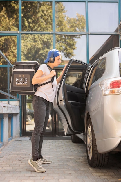 車で食品を配達するオフィス ランチ配達サービスの宅配便業者は、食品の入ったバックパックを保持しています。女, 聞くこと, へ, 音楽, 中に, ヘッドホン, キャリングバッグ, ∥で∥, 食事, 地位, 近くに, 自動車, サイド光景