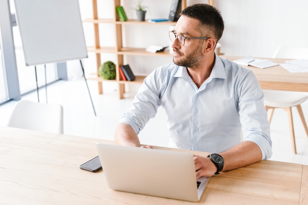 office man 30s in white shirt sitting at table and using laptop, while working in business centre