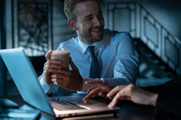 Office manager man drinking coffee smiling to his business partner laptop with cup of coffee in the office female hands typing on the keyboard Concept of overwork