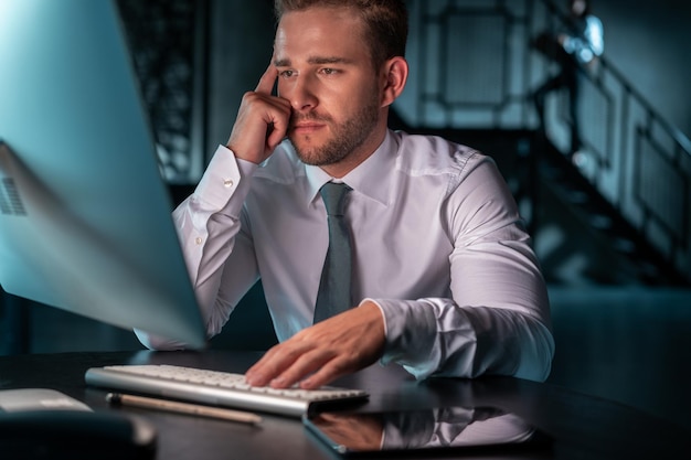 Office manager serious man thinking in front of computer\
monitor hand on the keyboard male manager alone in white shirt\
working in office late at night concept of overwork