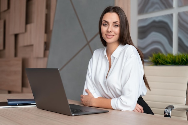 Office manager vrouw dragen wit overhemd glimlachend zittend aan tafel met laptop zijaanzicht kijken naar de camera Gelukkig kantoormedewerker in grote lichte kantoorruimte Concept van werk