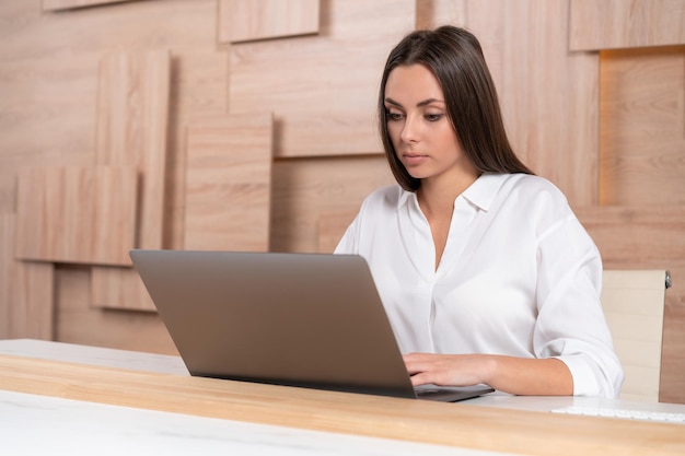 Office manager vrouw dragen wit overhemd zittend aan tafel met laptop op de achtergrond van houten kantoor muur zijaanzicht Geconcentreerde kantoormedewerker kijken naar het scherm Concept van werk