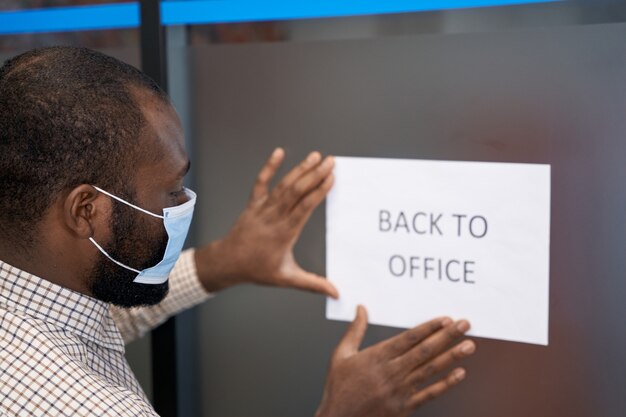 Office opening young afro american man in protective face mask sticking open sign with text back to