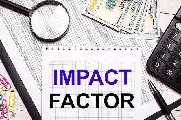 사무실 테이블에는 IMPACT FACTOR라는 텍스트가 있는 노트북이 있습니다. 펜 계산기 달러 여러 가지 빛깔의 종이 클립과 돋보기 세련된 직장 비즈니스 개념