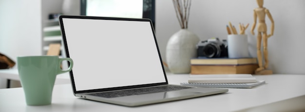 Office table with blank screen laptop, coffee mug, stationery and other supplies