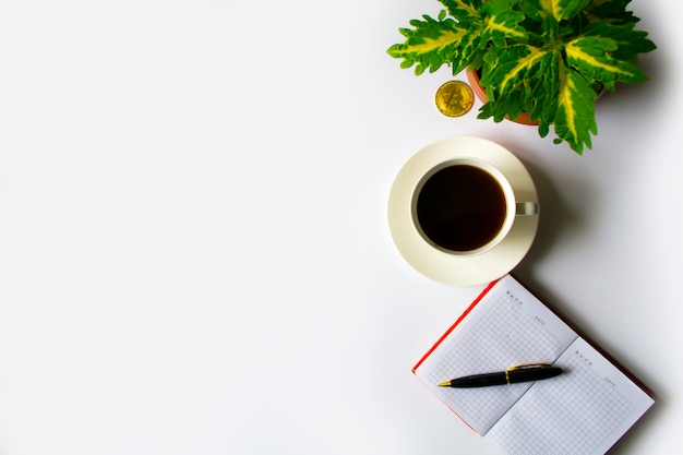 An office table with a cup of coffee next to a notebook in a red note nearby is a green flower and b...