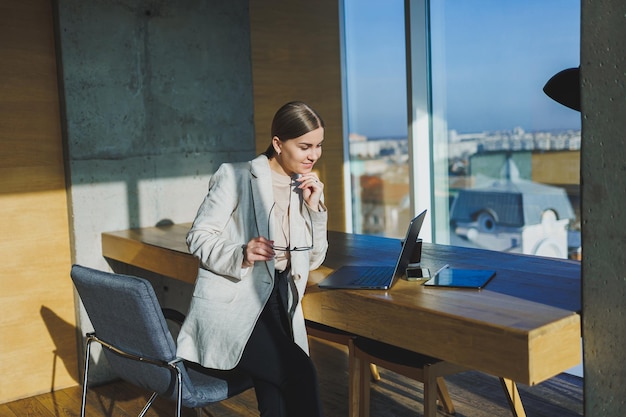 Office work attractive young female manager working in office standing in bright workspace using modern laptop computer young woman drinking delicious coffee