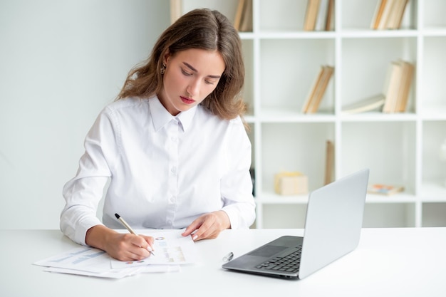 Office work Elegant woman Everyday routine Elegant concentrated lady in white shirt sitting desk opened laptop checking documents in light room interior