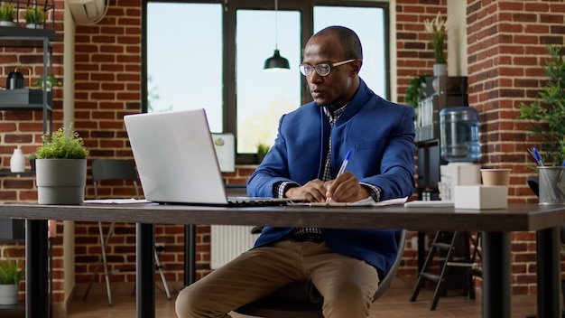 Office worker analyzing research information on laptop and taking notes to plan business strategy. Corporate employee working on project presentation with online statistics and paperwork.