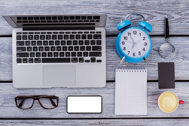 Office worker morning concept. Laptop with smarthphone, notepad, alarm clock and cup of coffee on a wooden table. Flat lay top view.