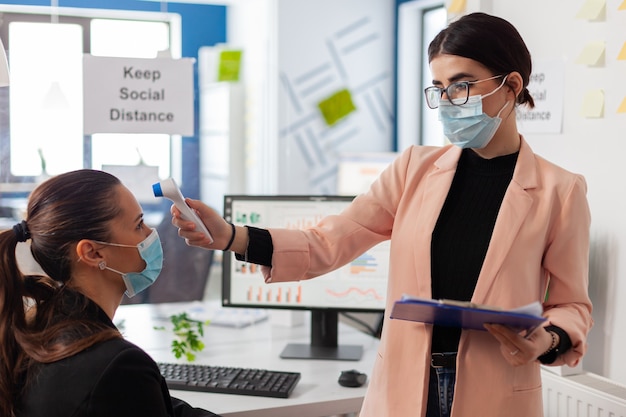 office worker scanning colleague temperature pointing digital thermometer at forhead keeping scoial distancing, during global pandemic with covid-19, wearing face mask as prevention.