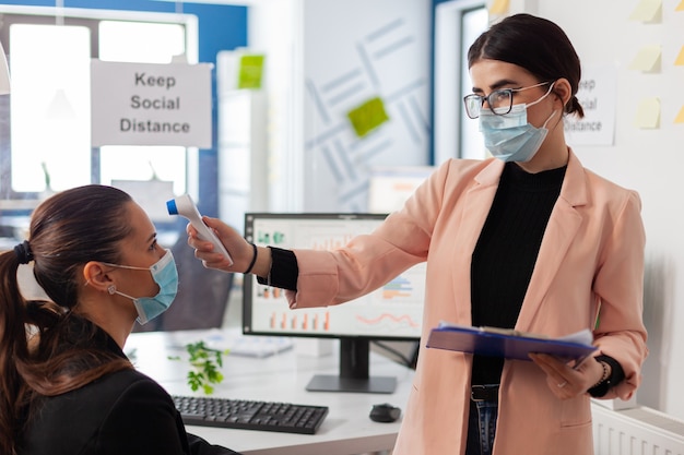 Office worker scanning colleague temperature pointing digital thermometer at forhead keeping scoial distancing, during global pandemic with covid-19, wearing face mask as prevention.