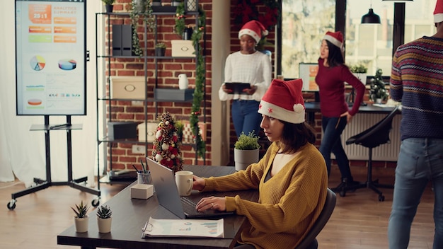 Office worker sending email on laptop during christmas holiday, doing startup work in space with xmas tree and decorations. Woman wearing santa hat at job in space with festive ornaments.