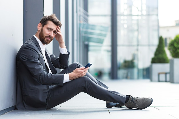 Office worker sitting near office outdoors got bad news, depressed and tired reads bad news from phone online