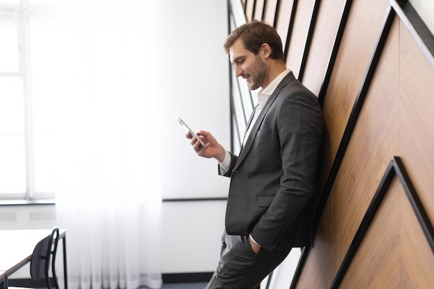 An office worker stands leaning against a wall and looks at a mobile phone