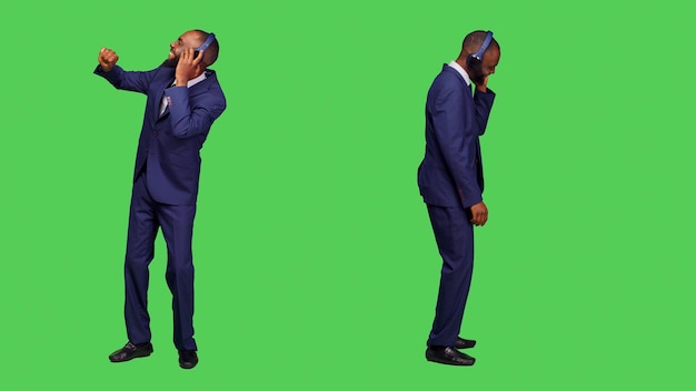 Office worker using headphones listening to music in studio, having fun with mp3 songs over full body greenscreen backdrop. Startup manager in suit dancing and wearing audio headset.
