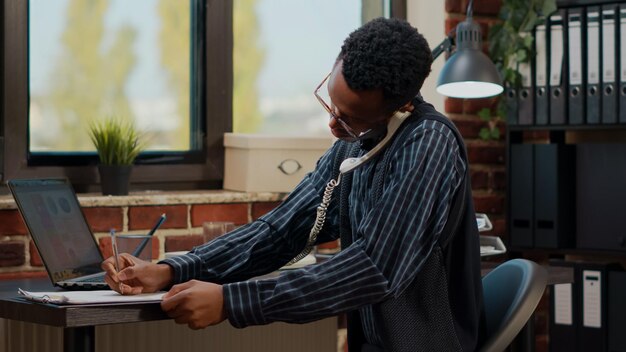 Office worker using landline phone to answer business call, discussing abput commercial strategy and innovation. Employee talking on telephone while he works on company development.