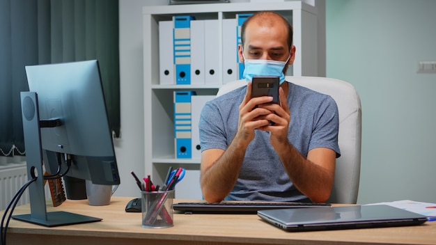 코로나바이러스 동안 현대 사무실에 앉아 전화 타이핑을 사용하여 보호 마스크를 쓴 회사원. 새로운 일반 직장에서 일하는 프리랜서 모바일 인터넷 기술을 사용하여 대화 쓰기