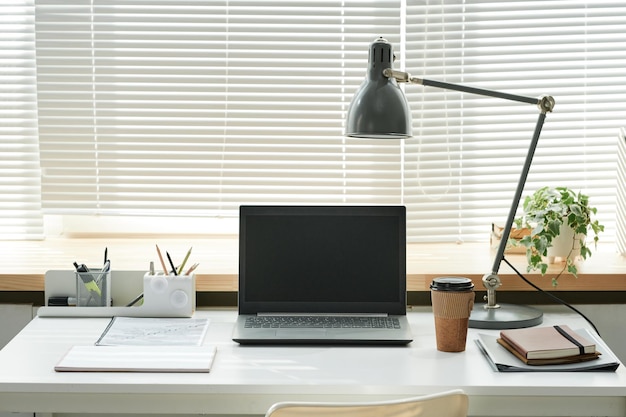 Office workplace with laptop lamp coffee cup and notepads against window closed with blinds