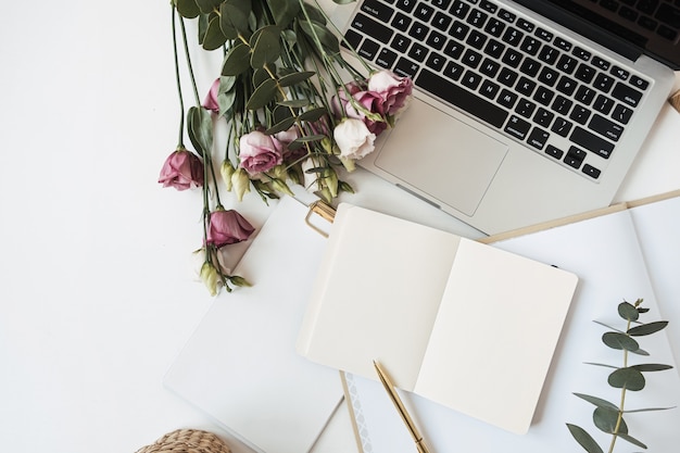 офисное рабочее пространство с блокнотом с чистым листом бумаги, ноутбуком, букетом цветов роз, веткой эвкалипта на белом столе