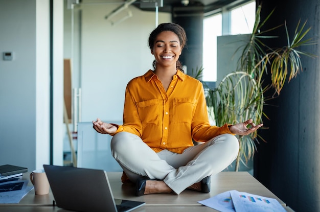 Офисный дзен Спокойная черная деловая женщина медитирует с закрытыми глазами на офисном столе, сидя на рабочем месте в позе лотоса