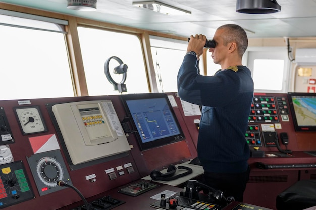 Officier op wacht met verrekijker op de navigatiebrug Blanke man in blauwe uniformtrui op de brug van een vrachtschip