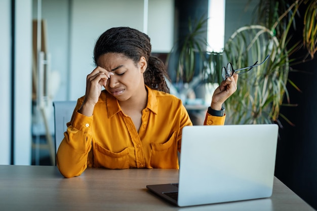Ogenvermoeidheid Vermoeide zwarte vrouwelijke kantoormanager die de neusbrug masseert en een bril vasthoudt terwijl hij op de werkplek zit