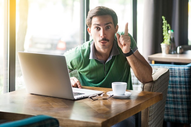 Oh I have an idea Young surprised businessman in green tshirt sitting with idea gesture and looking at camera with excited face business and freelancing concept indoor shot near window at daytime