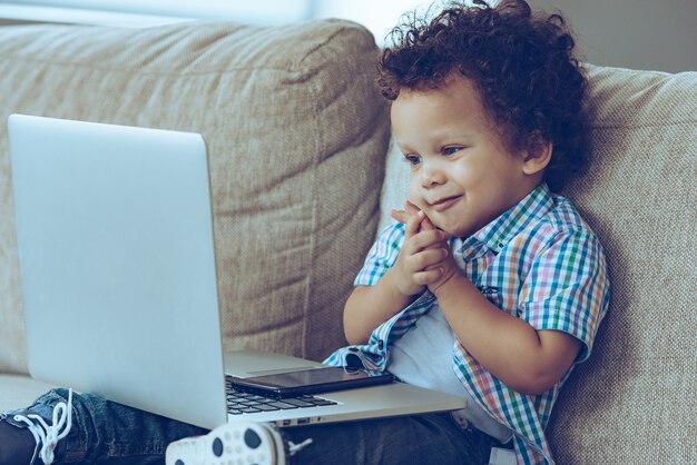 ああ、私のお気に入りの漫画をオンラインで！小さなアフリカの男の子は、自宅のソファに座って笑顔でノートパソコンを見ています。