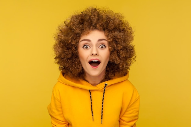 Oh my god, wow. Portrait of surprised curly-haired woman in urban style hoodie looking at camera with mouth open in amazement, expressing shock, astonishment. indoor studio shot, yellow background