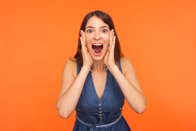 Oh my god wow Shocked impressed brunette woman in denim outfit standing with wide open mouth and clutching face in surprise looking startled amazed at camera indoor studio shot orange background