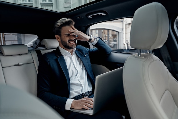 Oh no! Frustrated young man in full suit working using laptop and covering face with hand while sitting in the car