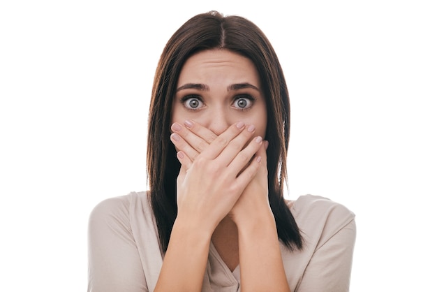 Oh no! Frustrated young woman covering mouth with hands and staring at camera while standing against white background