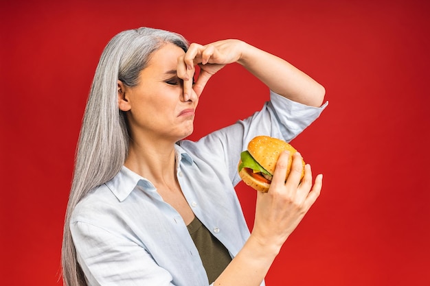 Oh così cattivo odore cibo stantio donna asiatica anziana matura che tiene hamburger nelle mani segnale di stop per cibo malsano isolato su sfondo rosso