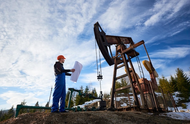 Operaio petrolifero in piedi nel giacimento di petrolio accanto a un martinetto della pompa con una grande carta