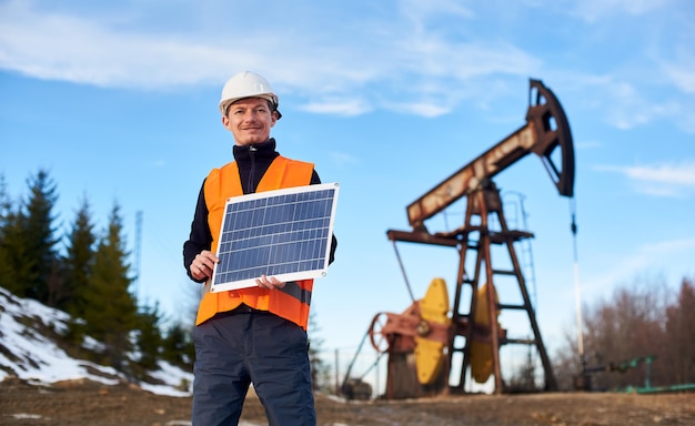 Oilman standing on an oilfield holding mini solar module next to an oil rig