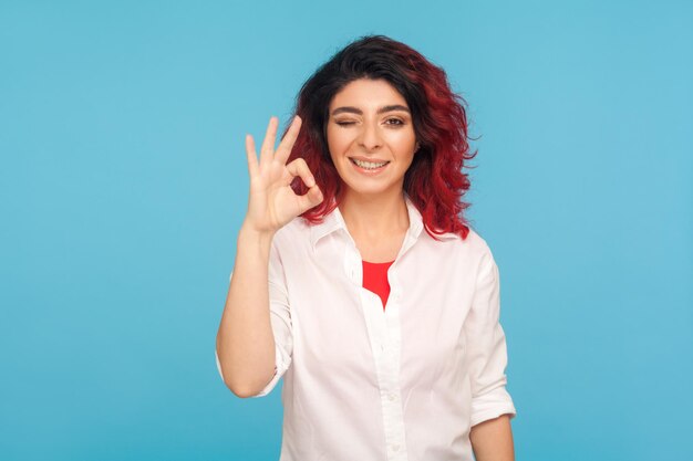 Oké, goedkeuringsteken. Portret van een gelukkige tevreden hipstervrouw met mooi rood haar in een wit overhemd dat knipoogt en een goed gebaar toont, alles is in orde. indoor studio-opname geïsoleerd op blauwe achtergrond