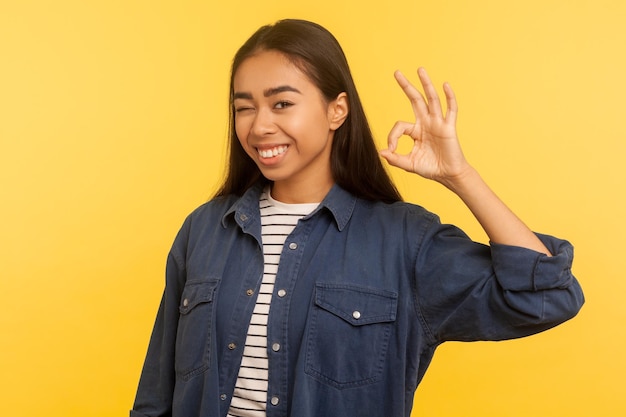 Oké, ik ben het ermee eens. Portret van een vrolijk lachend meisje in een spijkerblouse met een goed handgebaar, een cool symbool voor goedkeuring, tevreden met een uitstekend resultaat. indoor studio-opname geïsoleerd op gele achtergrond