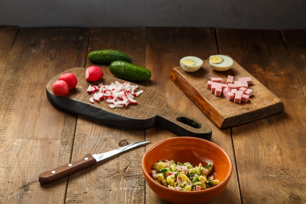 Okroshka in a clay plate on a table on a wooden table next to chopped vegetables and sausage on planks and a knife