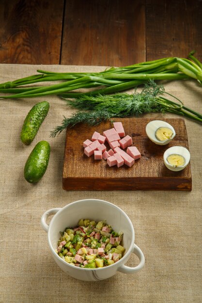 Okroshka in een soepkom op een houten tafel naast greens op planken en een mes