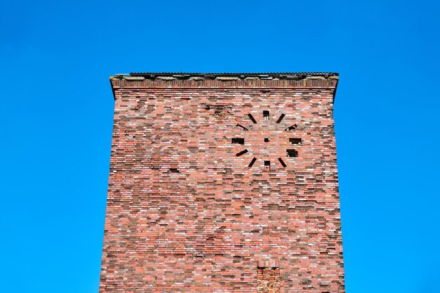 Vecchia torre abbandonata del mattone rosso con l'orologio rotondo sulla facciata contro il fondo del cielo blu, frammento. edificio industriale in muratura di mattoni a grana vintage, sfondo di architettura antica