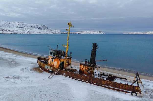 Старый заброшенный корабль на берегу Баренцева моря в Северном Ледовитом океане. Поселок Териберка Кольского полуострова. Разрушенная экономика и промышленность России, вид сверху с высоты птичьего полета