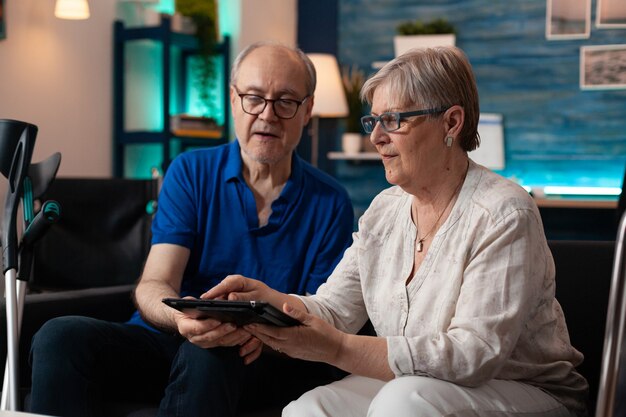 Old adult couple holding digital tablet at home on couch