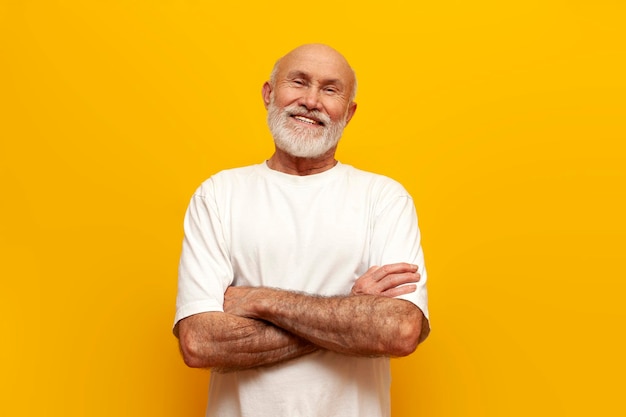 old bald man with gray beard in white Tshirt stands with his arms crossed on yellow isolated background and smiles