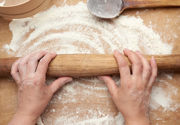 Old brown wooden rolling pin in female hands on a brown wooden space with white wheat flour
